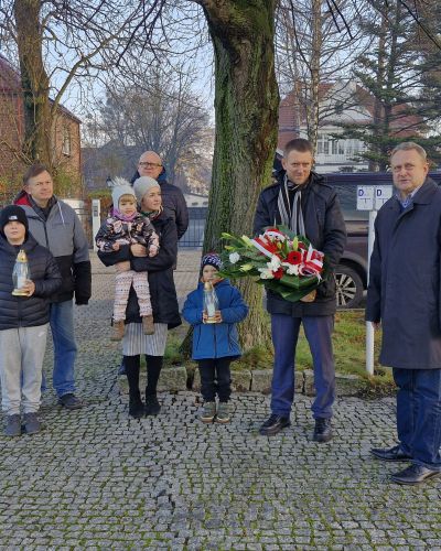 Grupa osób z dziećmi trzymających znicze i wieniec podczas uroczystego upamiętnienia